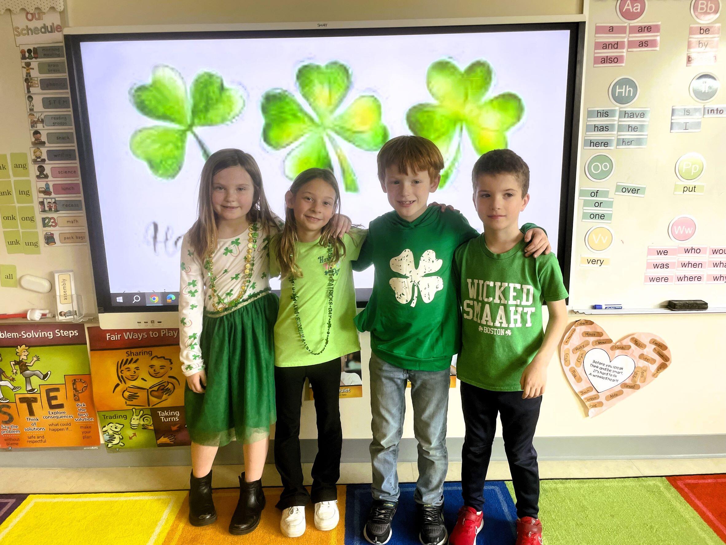 Enf-  Four students wearing green, standing in front of a smartboard. On the smartboard are shamrocks
