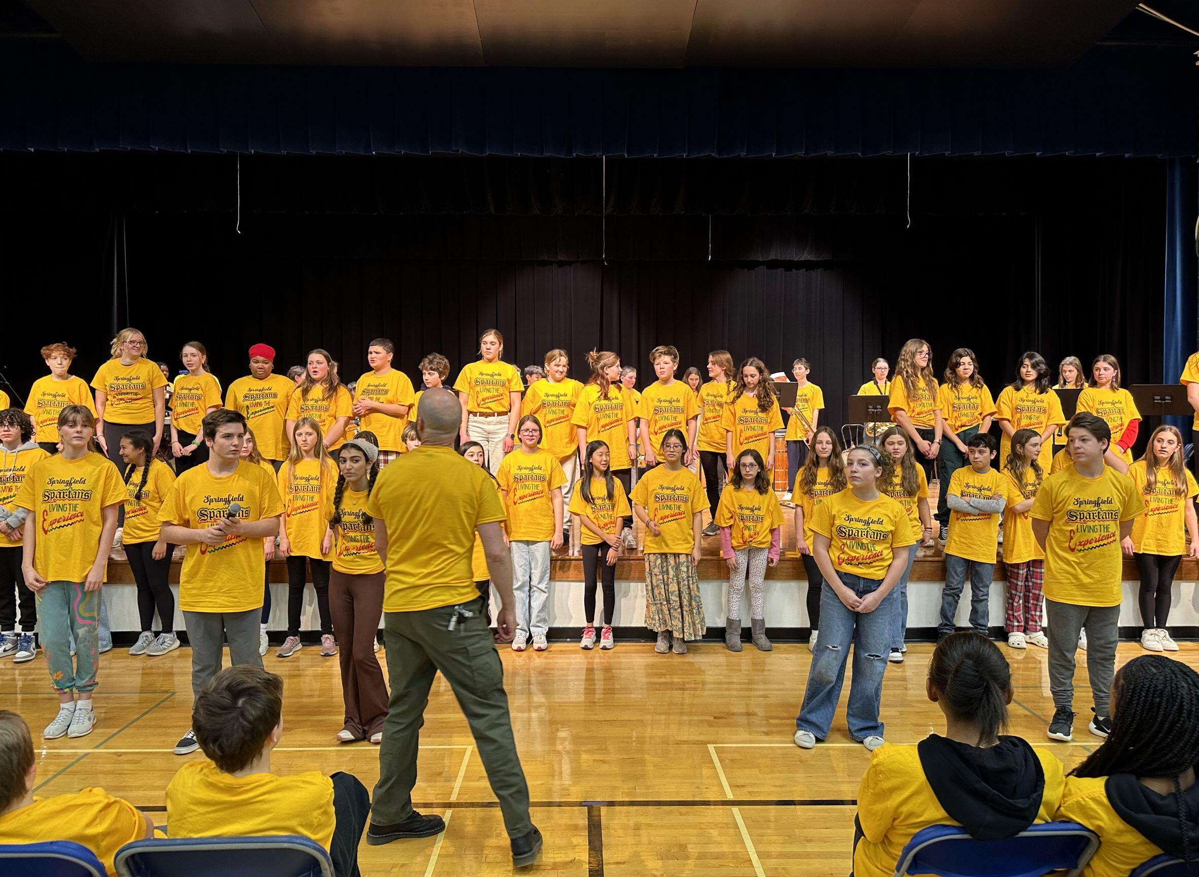 Chorus students practicing, all wearing the same yellow shirt that states: Springfield Spartans Living the Experience